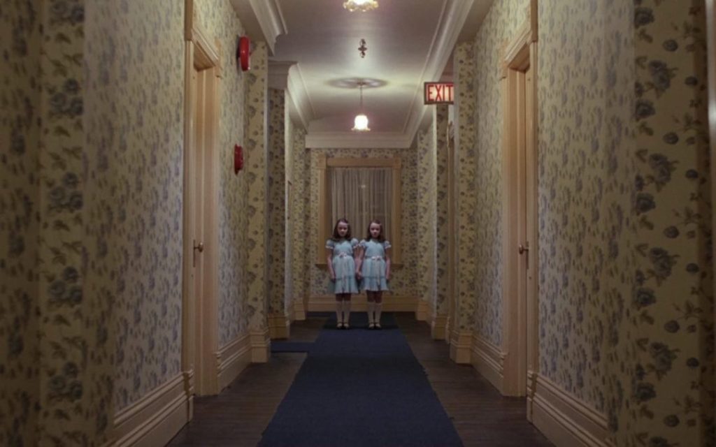 Two twin girls in matching dresses stand at the end of a hotel hallway