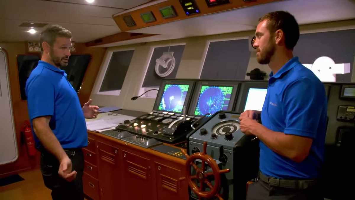 Two men talking to each other, wearing blue shirts, in a ship's bridge. 