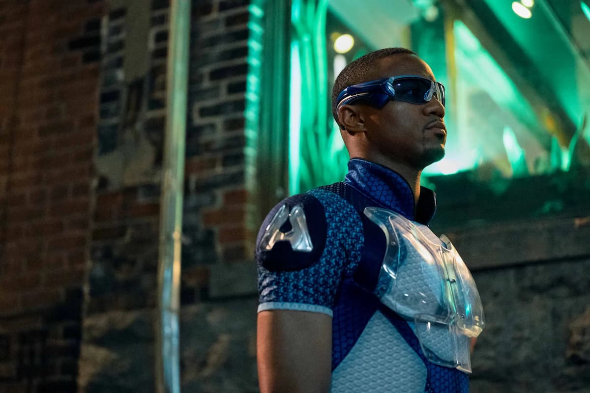 A-Train stands in an alleyway, staring off screen with a green window lit behind him