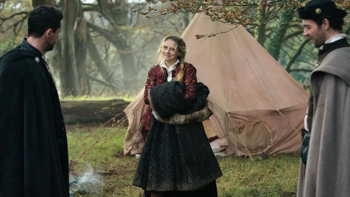 A Discovery of Witches - Diana and two men, Diana smiles at them as she holds a cloak outside in a forest 