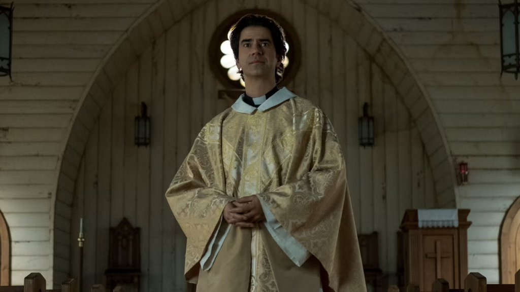 Hamish Linklater as Father Paul in Midnight Mass, standing in front of a church while wearing priestly regalia