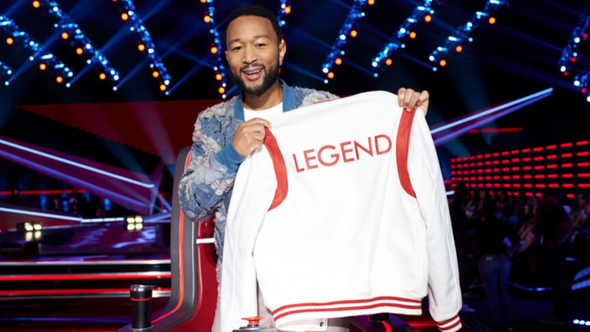 John Legend holding up a white jacket with the word "legend" embroidered on the back.