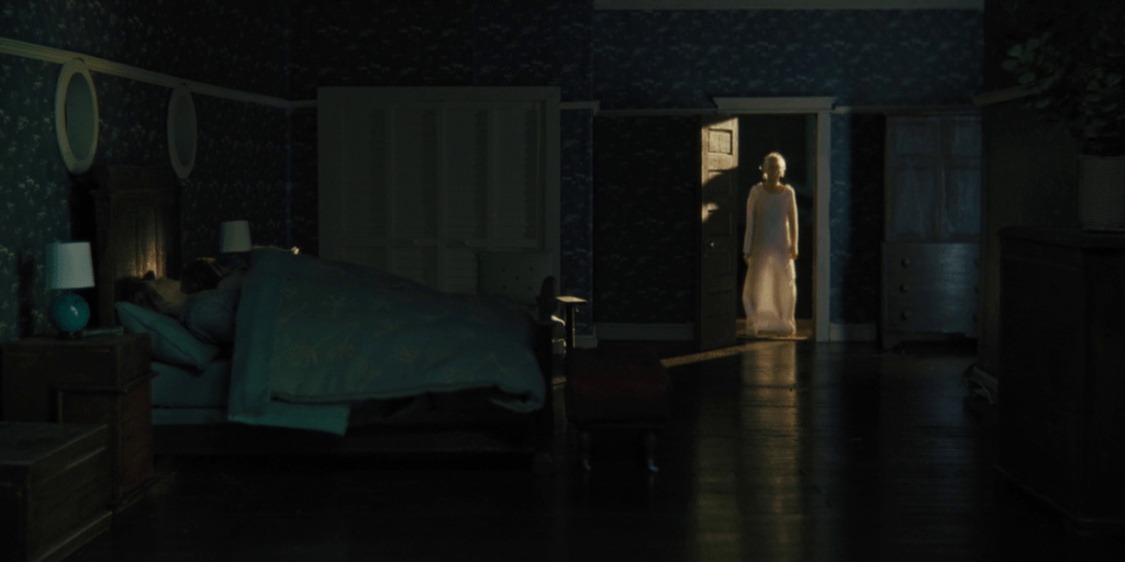 A woman stands in a white dress in the doorway of a bedroom 