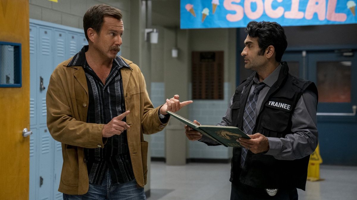 A man in a tan jacket speaks to a man in a vest with the word 'TRAINEE' on it in a highschool hallway