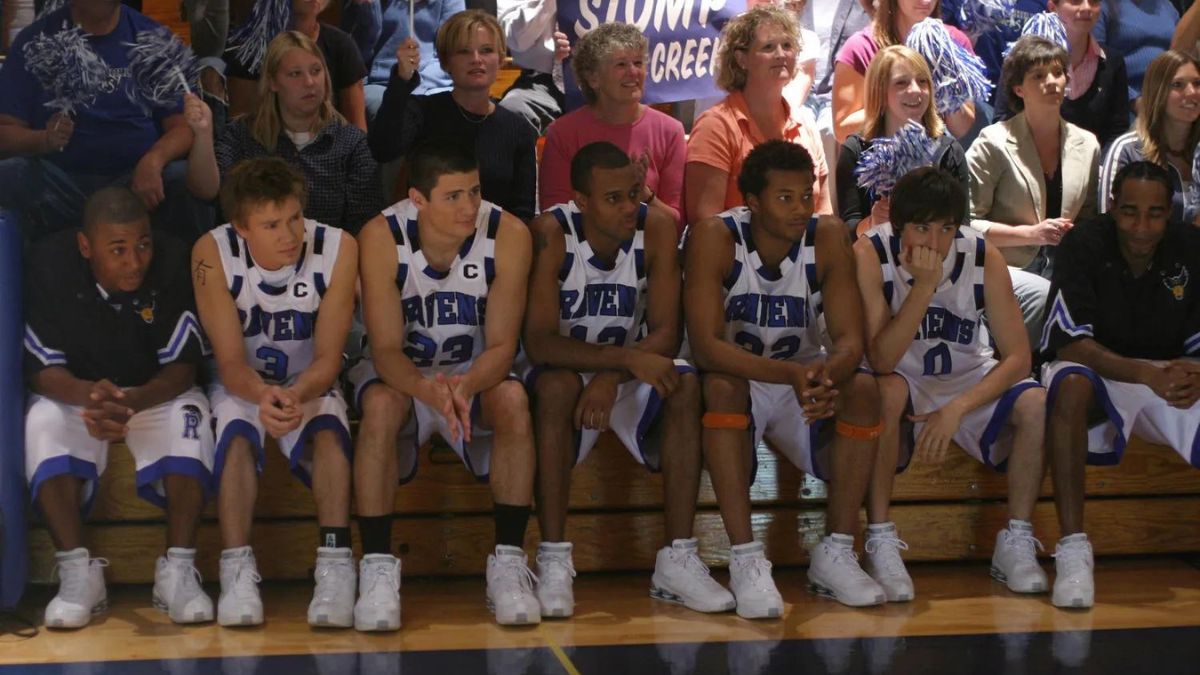 Several basketball players sit on a bench  beside a basketball court in One Tree Hill, a crowd of people behind them