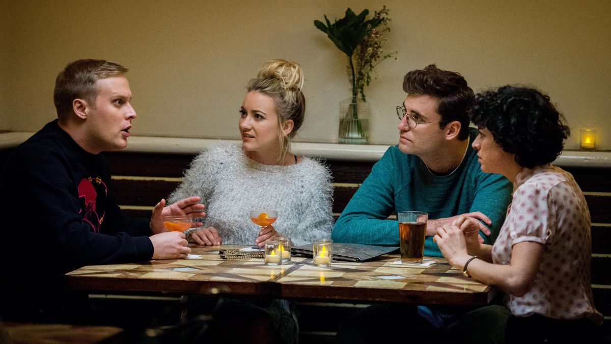 Four people sit around a table, two of them with cocktails in hand. The man at the end of the table animatedly speaks while the rest look at him.