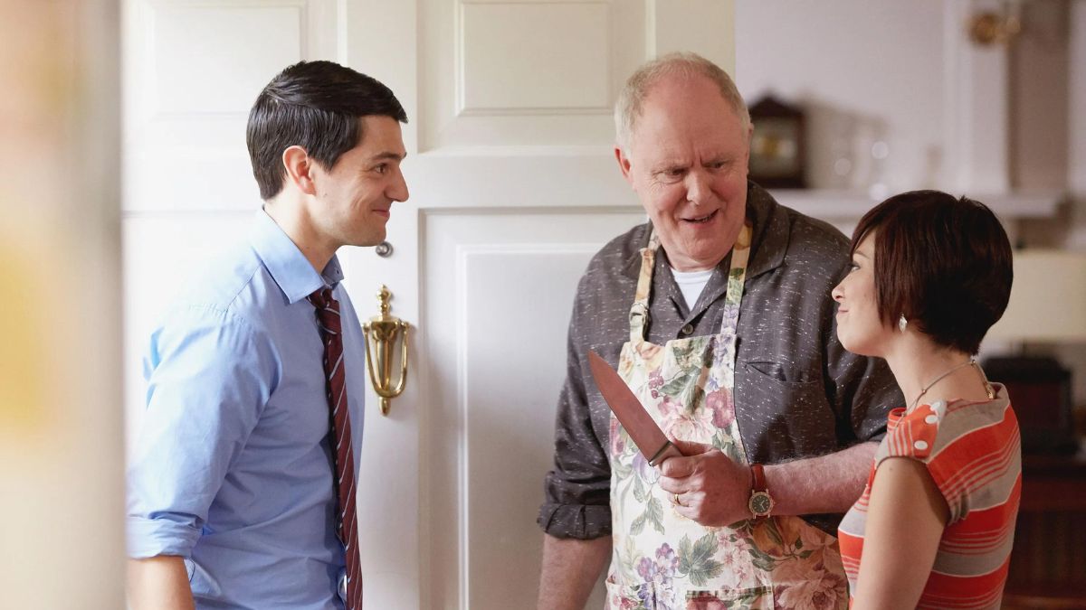 A man stands at the door of a house and two people greet him. One is an older man with an apron on and a knife, smiling down at a younger woman who smiles politely at the man at the door