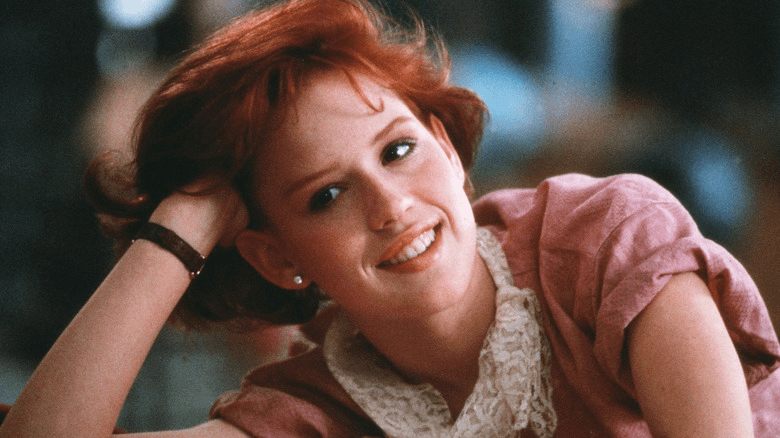 Molly Ringwald in The Breakfast Club, wearing a pink dress and leaning on one hand while she rests at a desk.