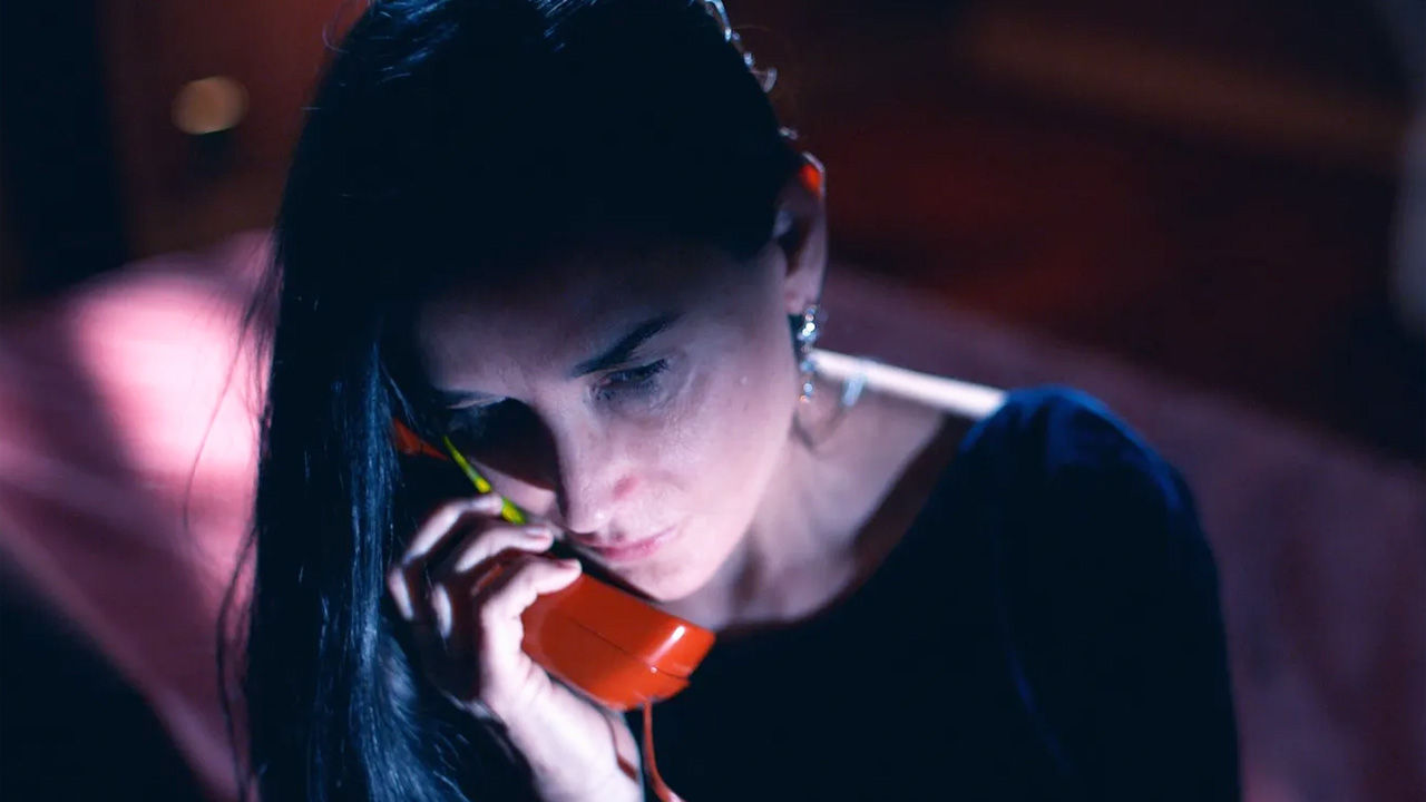 The Substance, Demi Moore's character talking into a red telephone, in a darkened room.