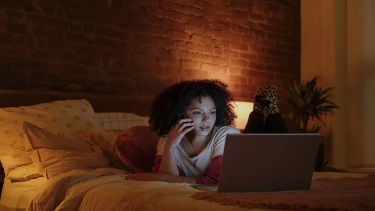 A woman laying on her bed talking on her phone while using a laptop.